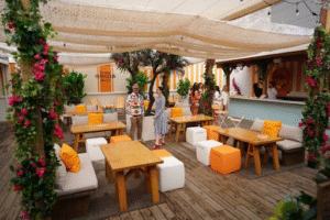 Top Rooftop Patios in Toronto. Soluna Toronto. Outdoor lounge area with wooden tables, cushioned benches, and orange-and-white cube stools under a canopy with hanging plants and flowers. People are standing and chatting near the bar in the background.